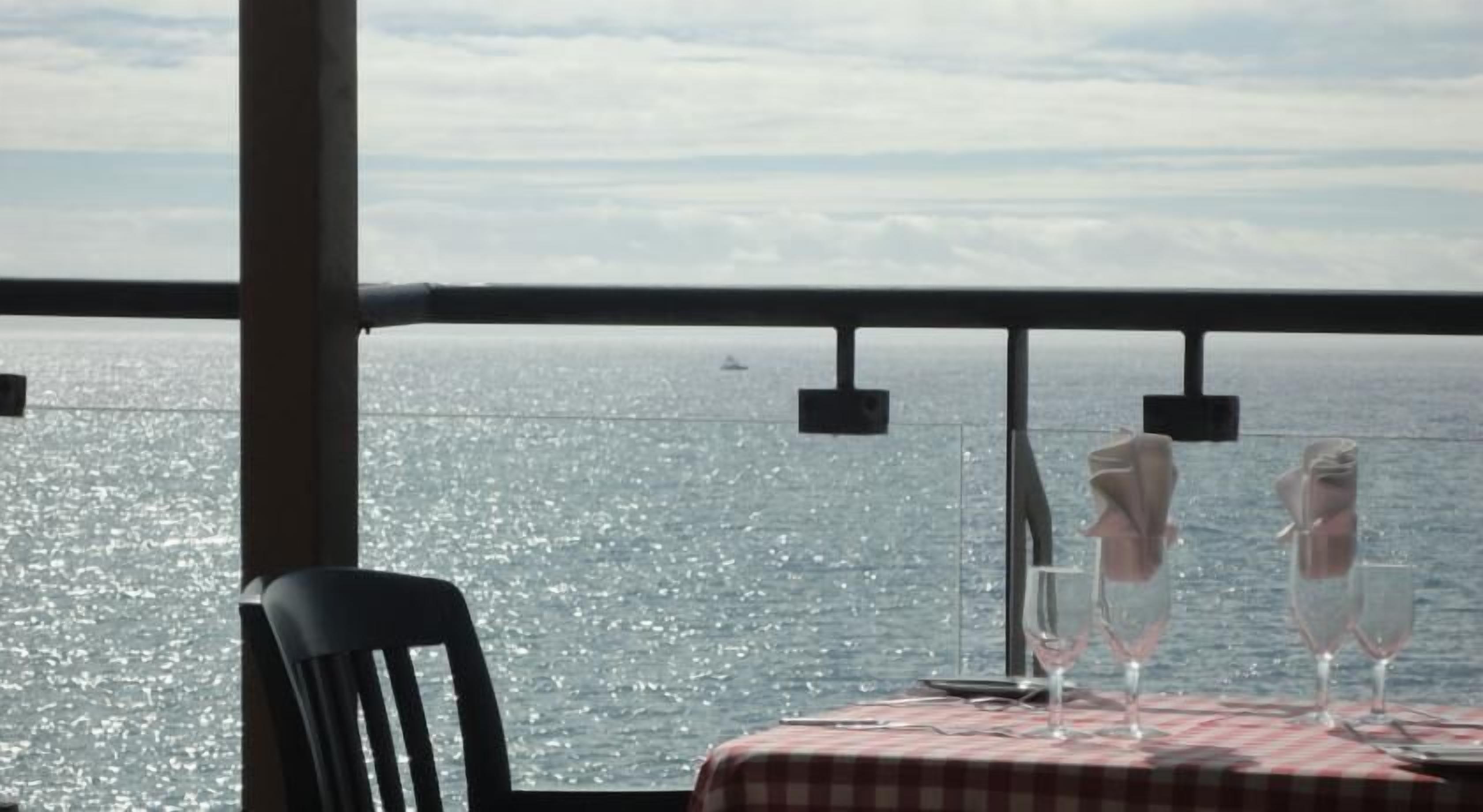 Una mesa con mantel a cuadros, dos copas de vino y una silla frente a una vista del mar.