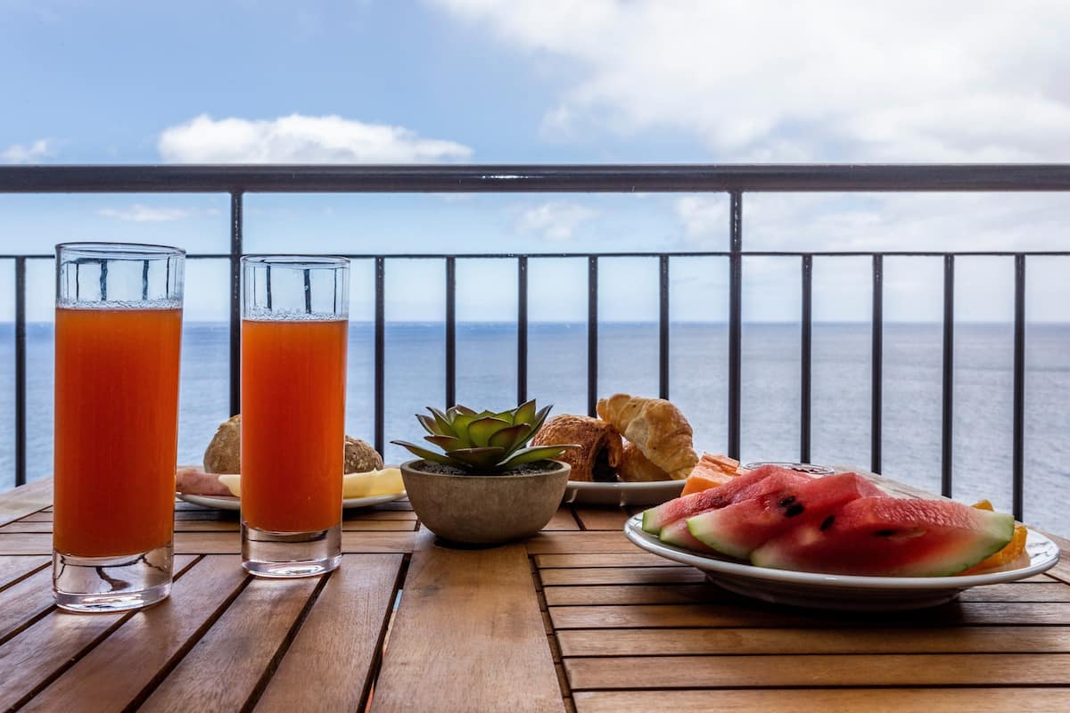 A wooden table with two glasses of orange juice, a plate of watermelon slices, a small plant, and some pastries on a balcony with a view of the sea.