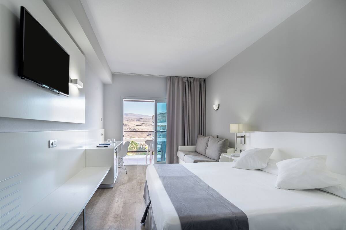 A modern hotel room with a bed, a desk, a TV, and a balcony with a view of the mountains.