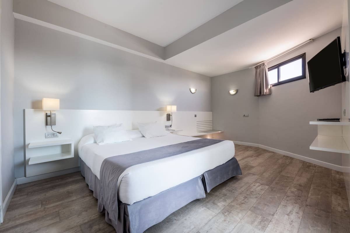 A modern bedroom with a large bed, bedside tables, a flat-screen TV, and a window with curtains.