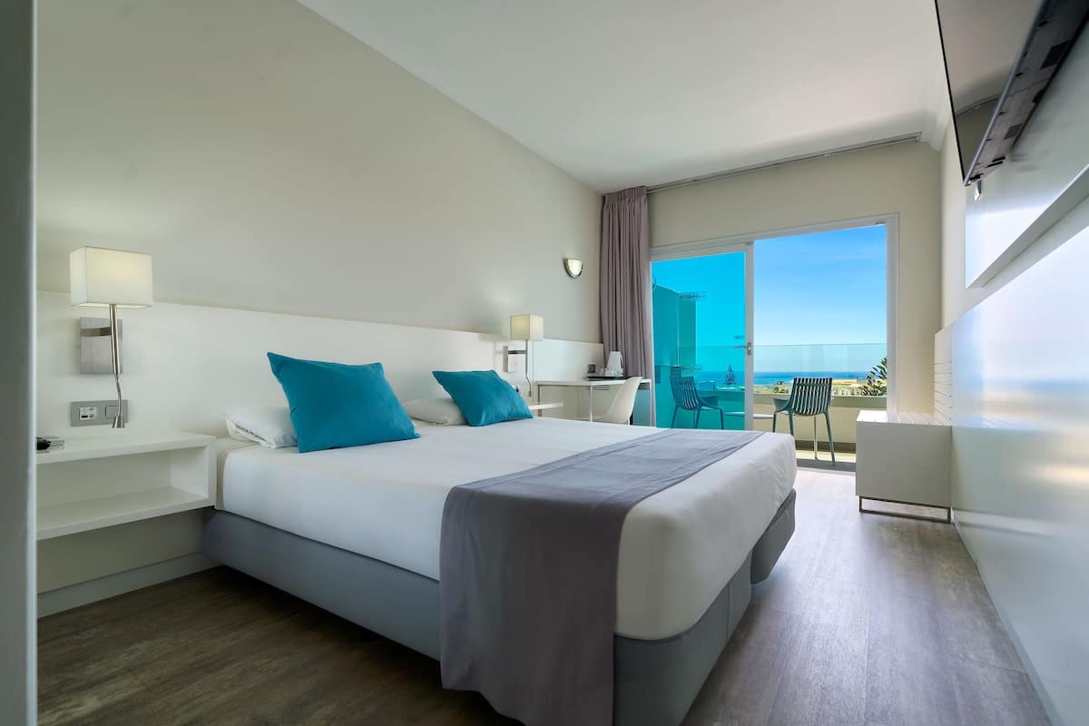 A modern hotel room with a large bed, two bedside tables, a desk, and a balcony with a view of the ocean.