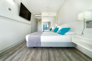 Una habitación de hotel moderna con una cama grande, un televisor de pantalla plana en la pared, una mesita de noche con lámpara y un espejo amplio.