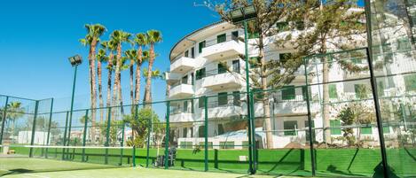 A tennis court with a net, surrounded by a green fence and palm trees.
