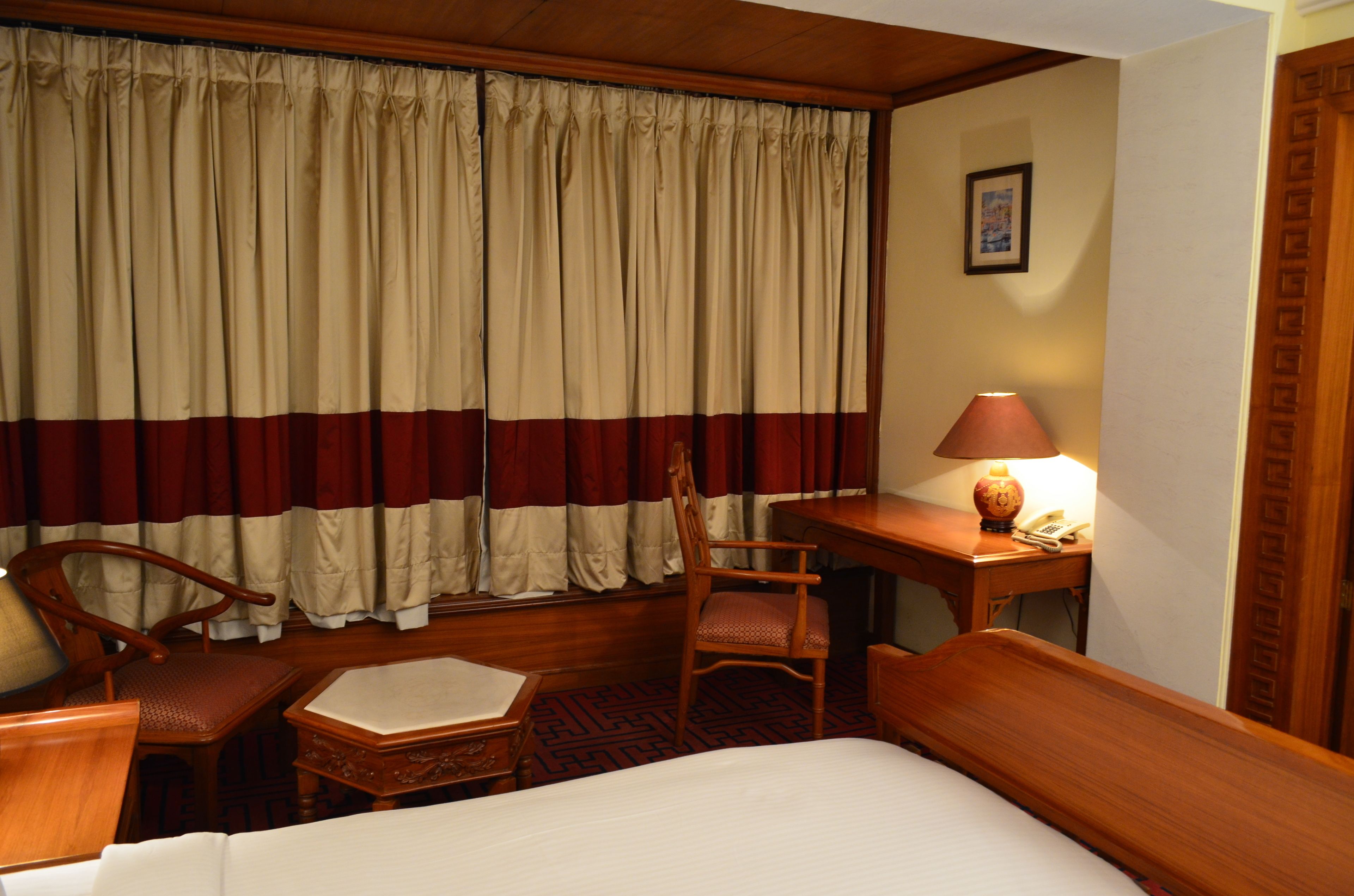 A hotel room with a bed, a desk with a lamp, a chair, and a window with curtains.
