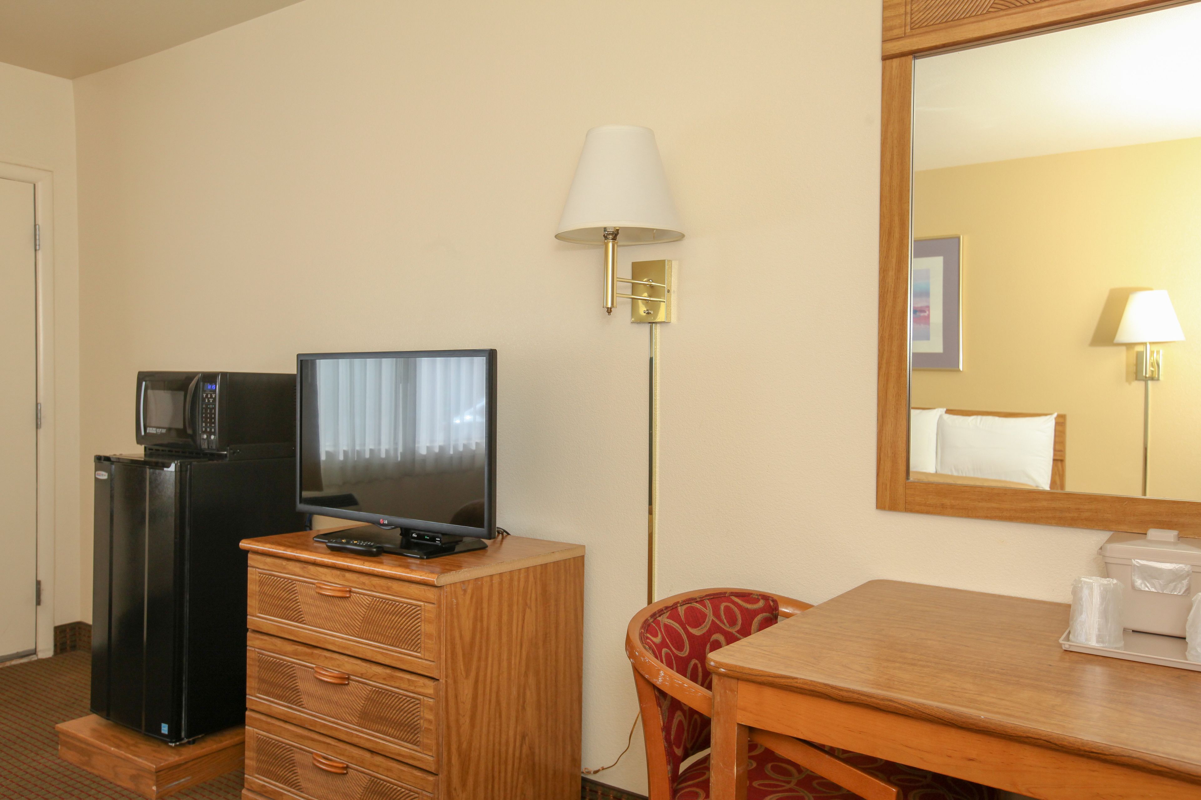 Habitación de hotel con televisor de pantalla plana, refrigerador, cómoda de madera, una silla, una mesa y un espejo.