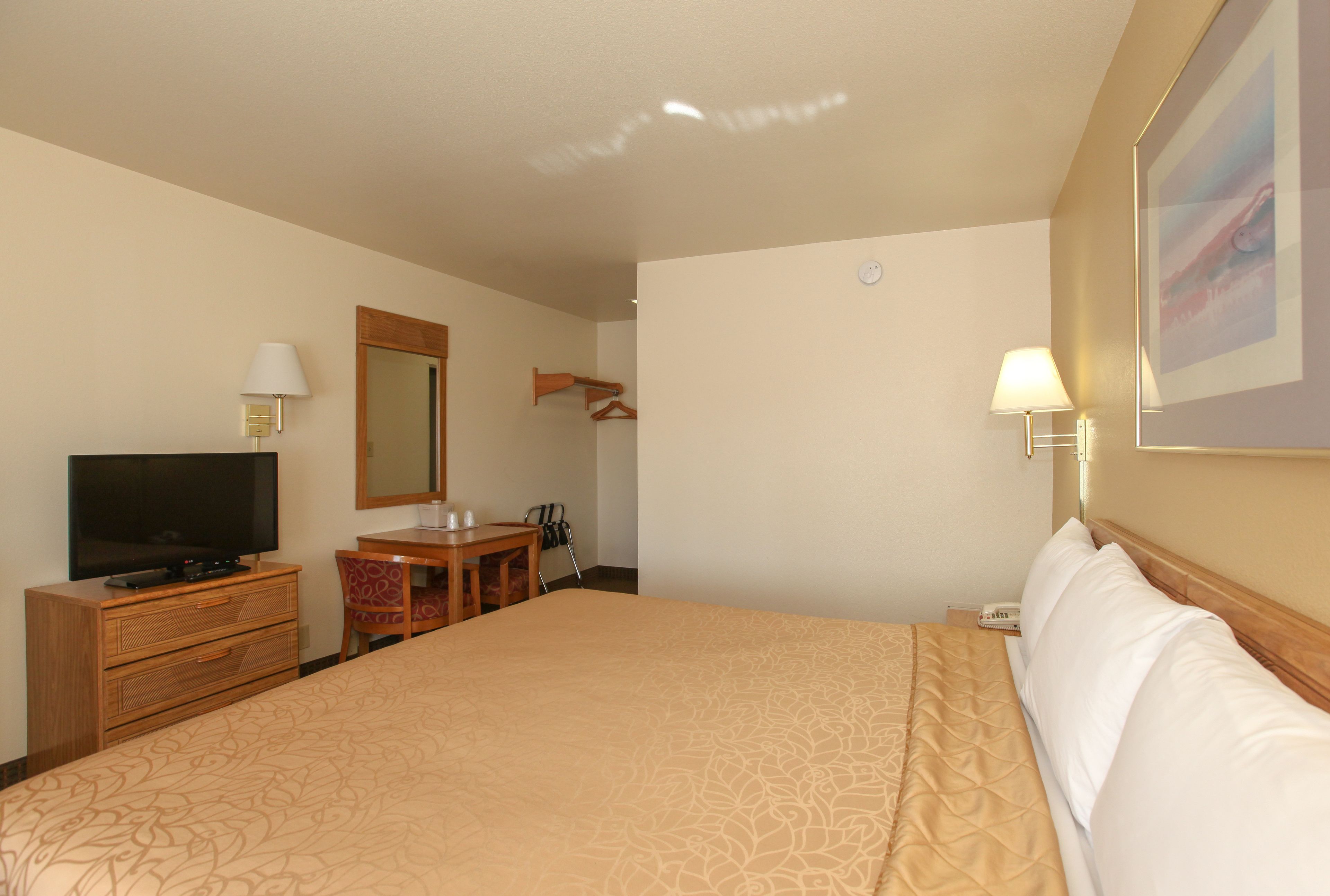 A hotel room with a large bed, a television, a desk with a chair, a mirror, and a wall-mounted lamp.