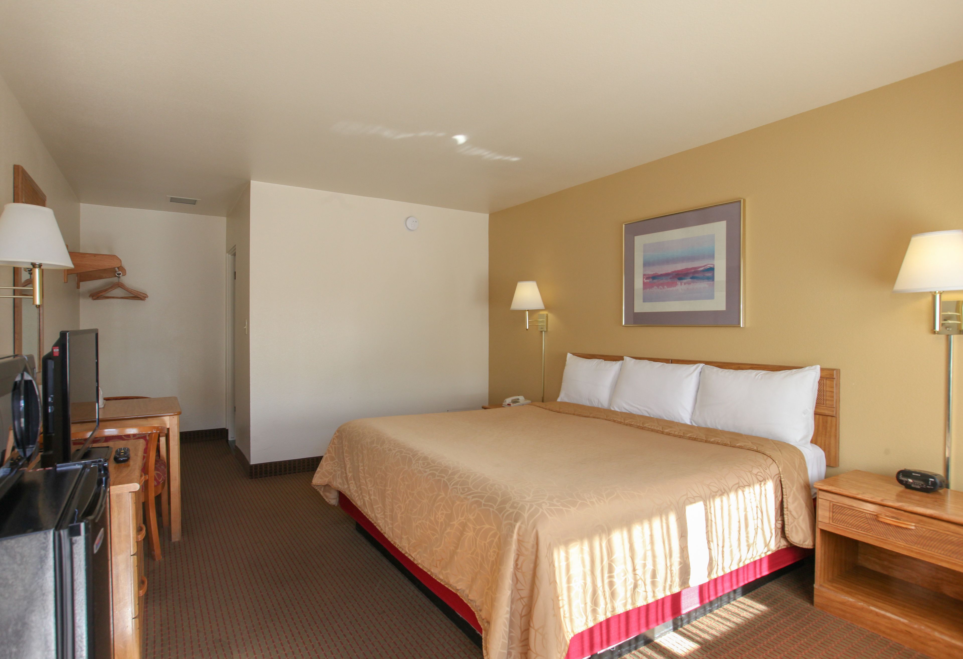 Quarto de hotel com cama, mesas de cabeceira, escrivaninha com computador, televisão e cabideiro.