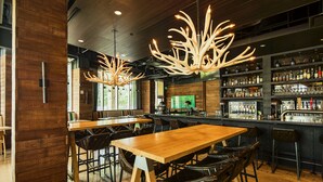 Un área de bar con mesas y sillas de madera, una barra con estantes llenos de botellas y una gran lámpara de araña.