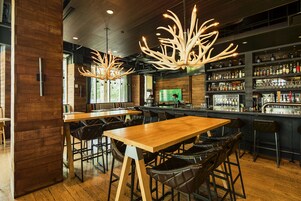 A bar area with wooden tables and chairs, a bar counter with shelves of bottles, and a large chandelier.