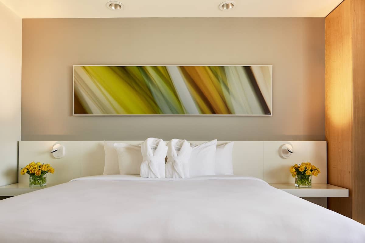 A modern hotel room with a large bed, two nightstands, a wall-mounted artwork, and a vase of flowers.