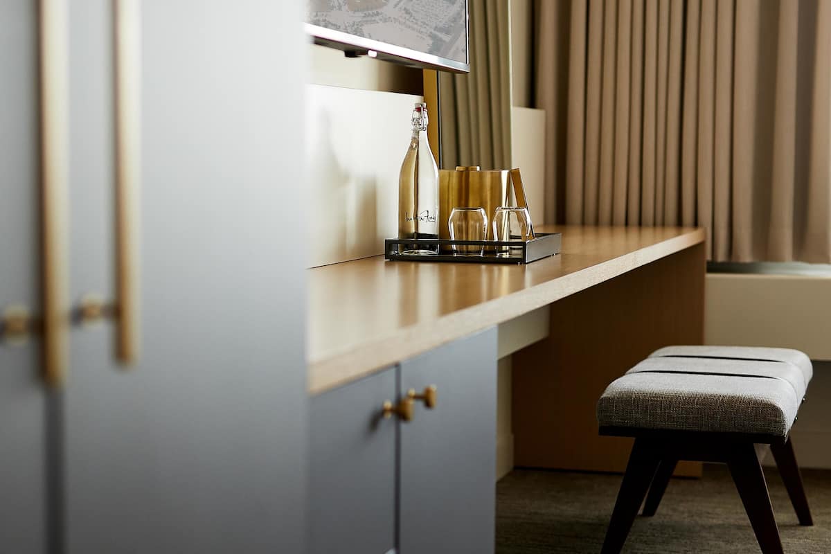 A modern hotel room with a wooden desk, a framed picture, a bottle, glasses, and a bench.