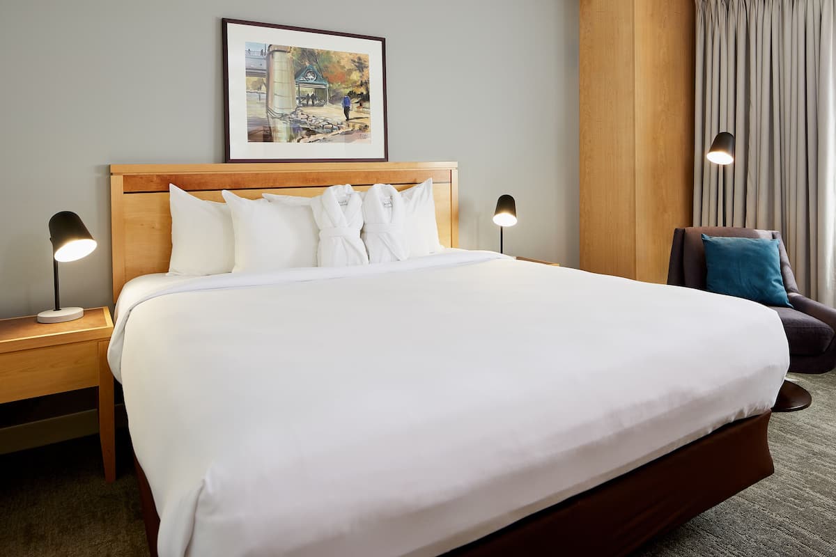 A neatly made bed with white linens, two bedside tables with lamps, a framed picture on the wall, and a chair with a cushion.