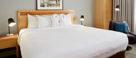 A neatly made bed with white linens, two bedside tables with lamps, a framed picture on the wall, and a chair with a cushion.