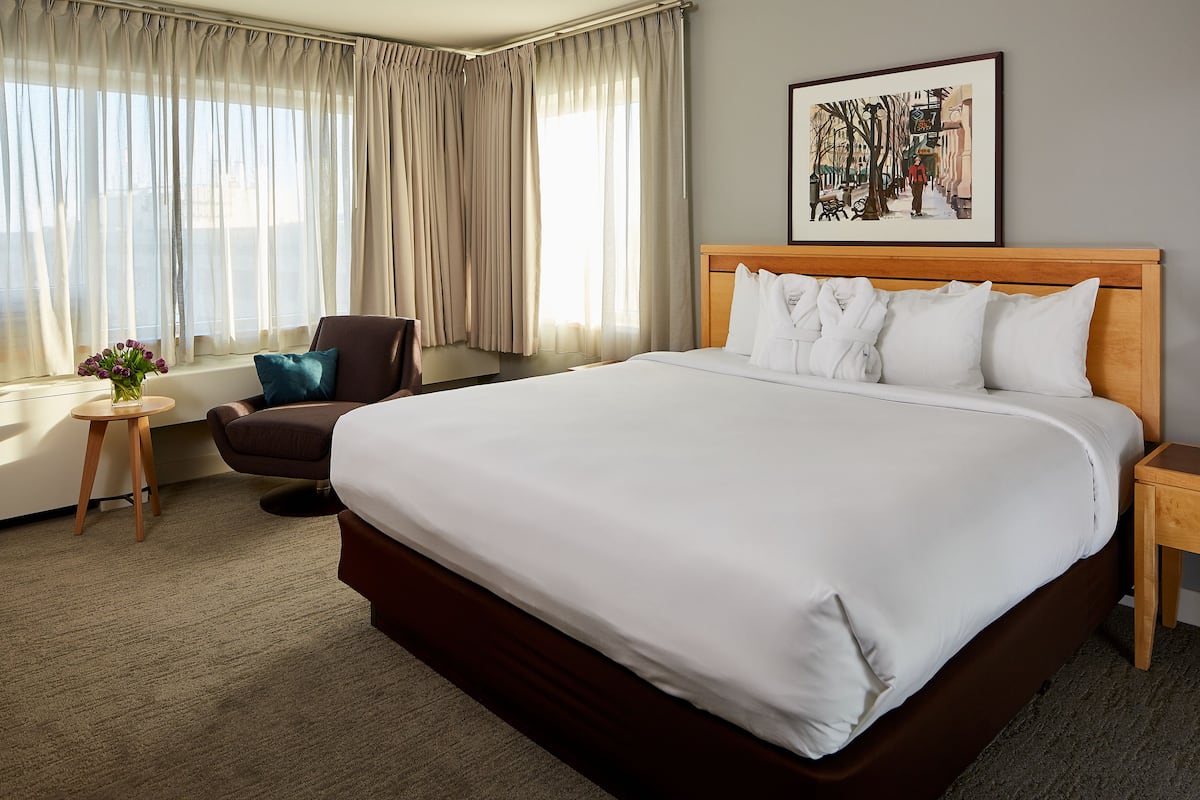 A hotel room with a large bed, a chair, a small table with a vase, and a painting on the wall.