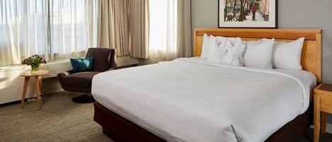 Una habitación de hotel con una cama grande, una silla, una mesita con un florero y un cuadro en la pared.
