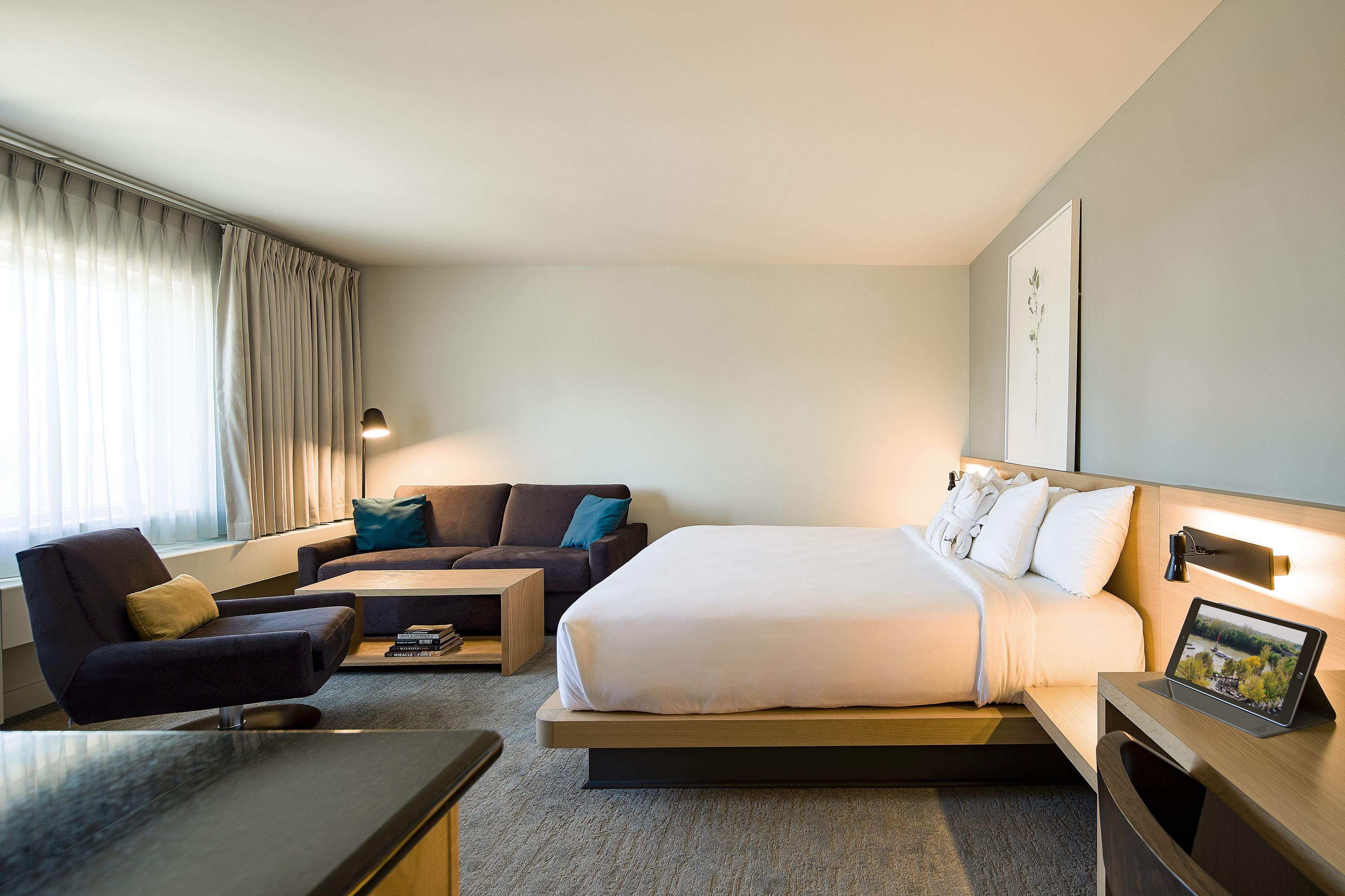 A modern hotel room with a bed, a sofa, a coffee table, and a desk with a tablet.