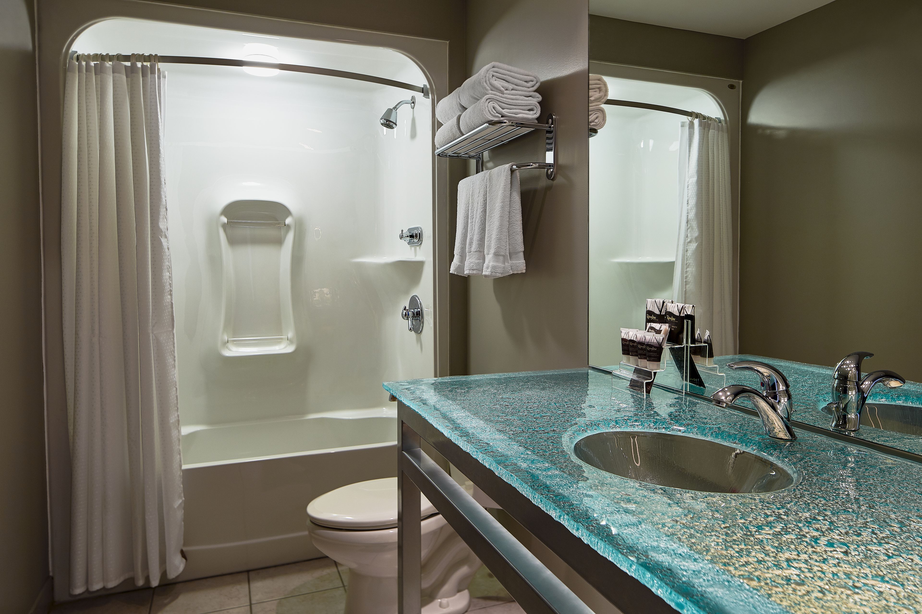 A bathroom with a glass shower enclosure, a white toilet, a sink with a glass countertop, and a mirror.