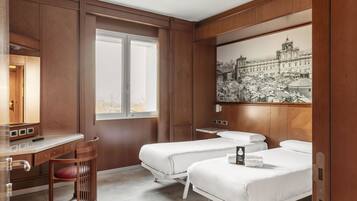 Habitación de hotel con un escritorio de madera, dos camas, una silla, un ventanal grande y una pintura de paisaje urbano en la pared.