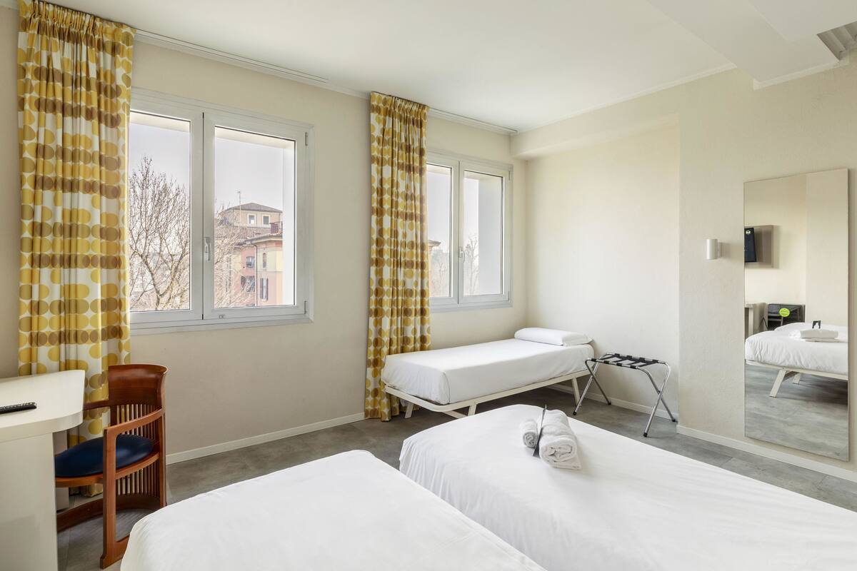 Quarto de hotel com duas camas, uma escrivaninha, uma cadeira, uma mesinha e uma janela com cortinas.