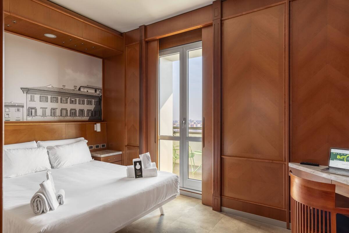 Quarto de hotel com cama, escrivaninha, laptop e vista para edifícios através de uma porta de vidro.
