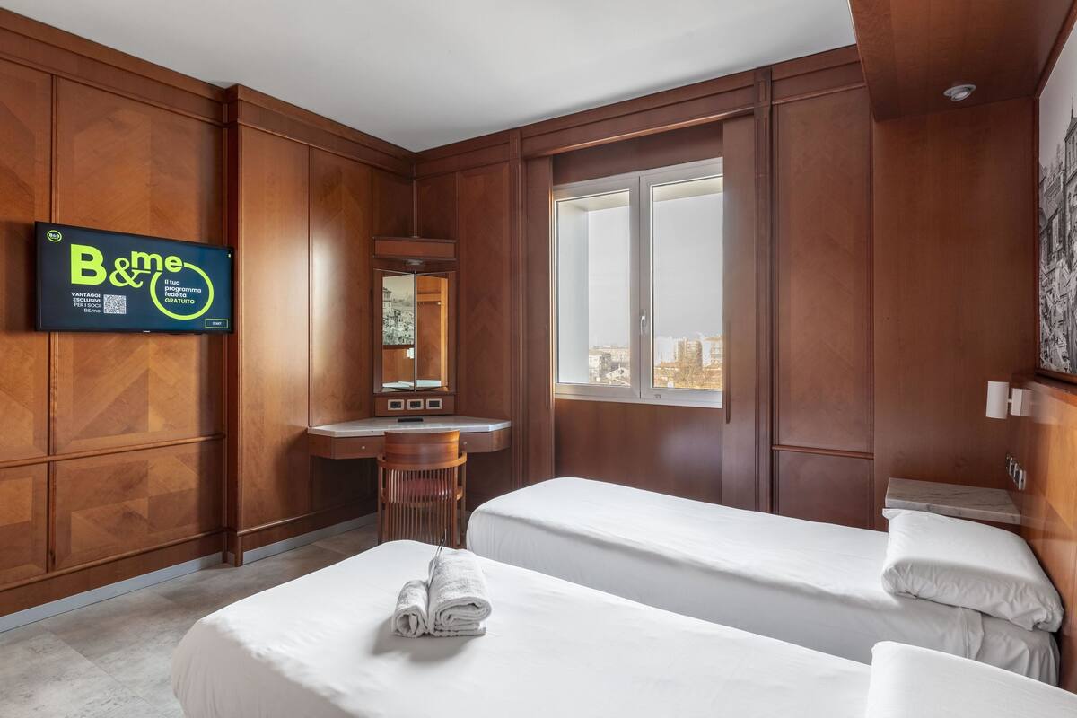 Quarto de hotel com duas camas, uma escrivaninha, um espelho e uma janela grande.