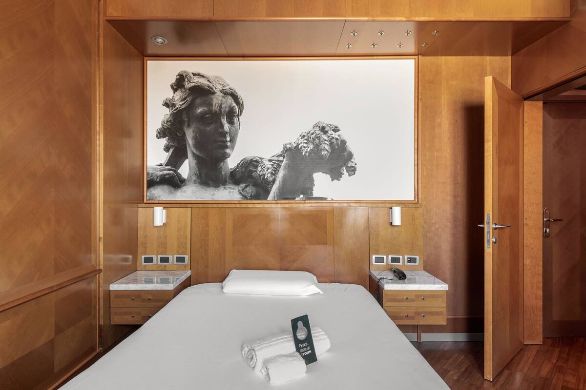 Quarto de hotel com uma cama grande, cabeceira de madeira, uma obra de arte na parede e uma pequena mesa de cabeceira com um telefone e um abajur.