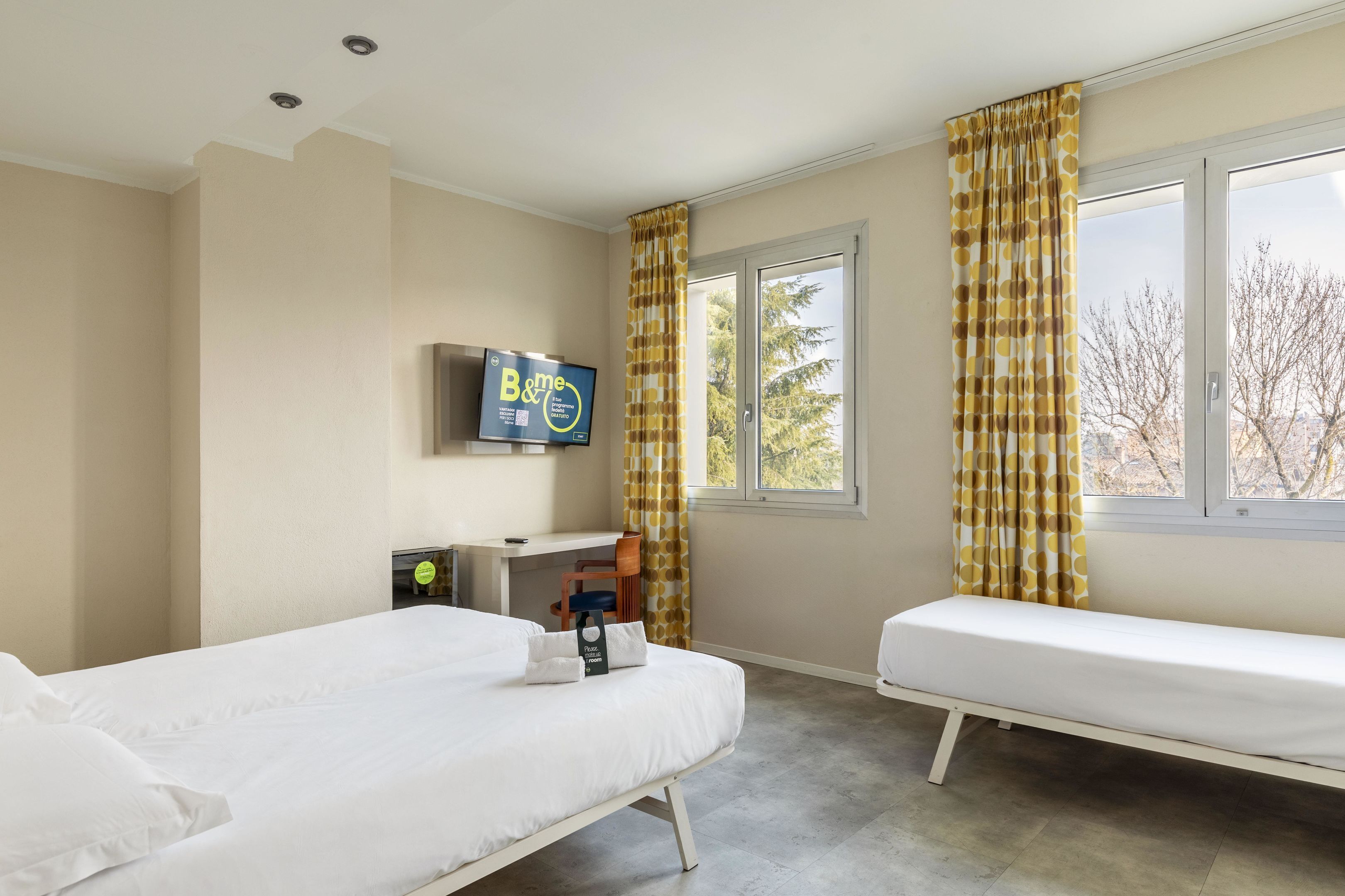Habitación de hotel con dos camas, un escritorio, un televisor y ventanas grandes con cortinas.