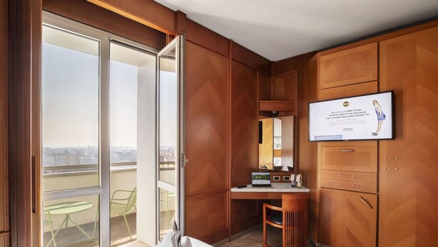 Une chambre d’hôtel dotée d’une grande porte-fenêtre coulissante donnant sur un balcon, d’un bureau avec un ordinateur, d’un téléviseur fixé au mur et de meubles en bois.