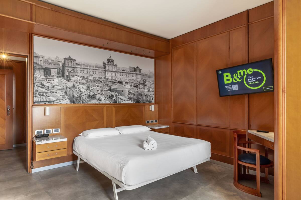 Quarto de hotel com uma cama grande, um guarda-roupa de madeira, uma escrivaninha e uma televisão exibindo um programa.