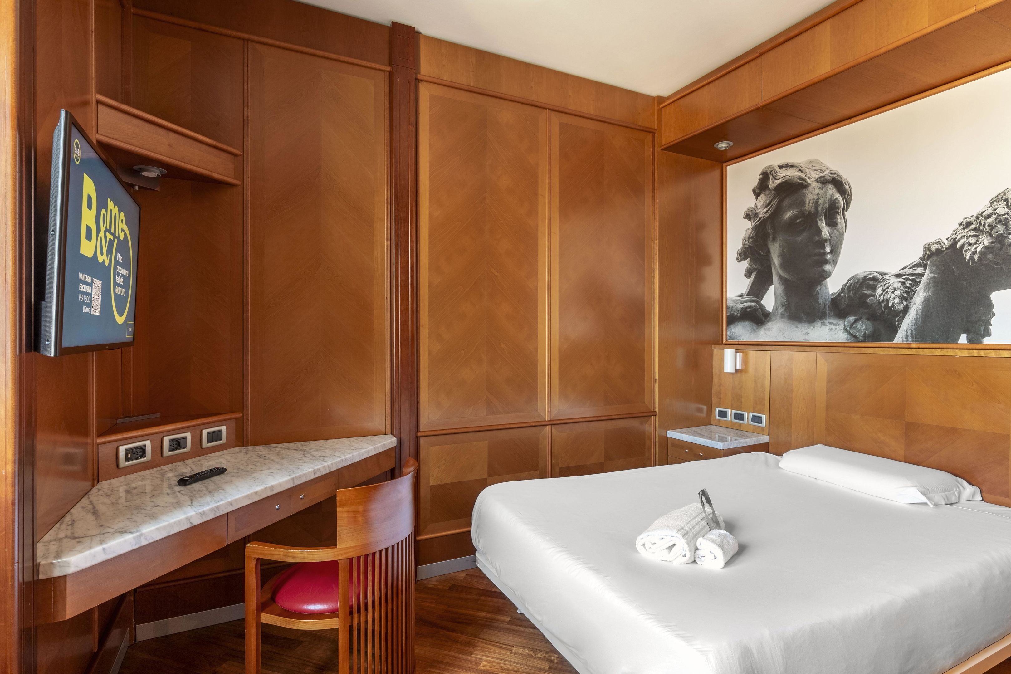 Ein Hotelzimmer mit einer Wandpaneele aus Holz, einem Marmorschreibtisch, einem Bett mit weißer Bettwäsche und einem großen Kunstwerk darüber.