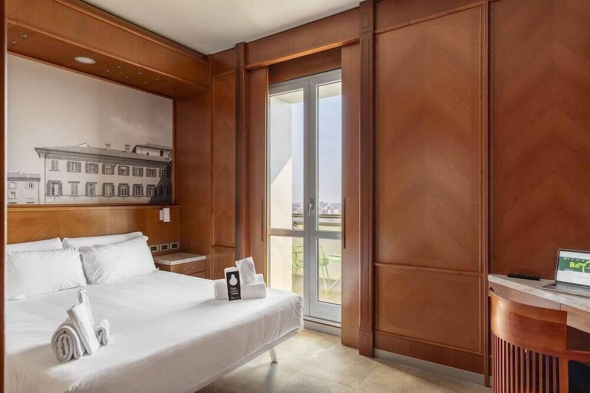 Quarto de hotel com cama, escrivaninha, laptop, janela com vista e obra de arte na parede.