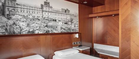 Habitación de hotel con dos camas, un gran mural de paisaje urbano en la pared, paneles de madera, una mesita con una toalla y una mesita de noche con una lámpara.