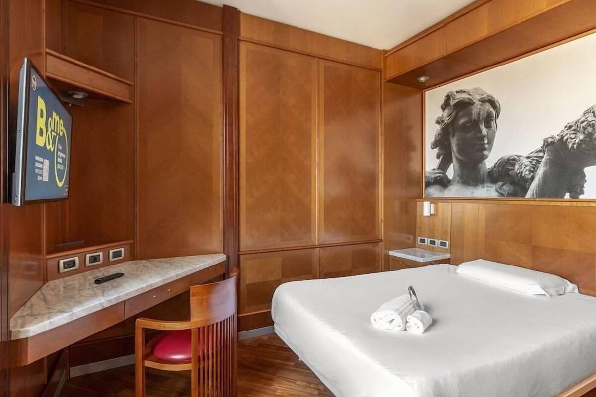 Quarto de hotel com cama, escrivaninha, cadeira, televisão e um quadro na parede.