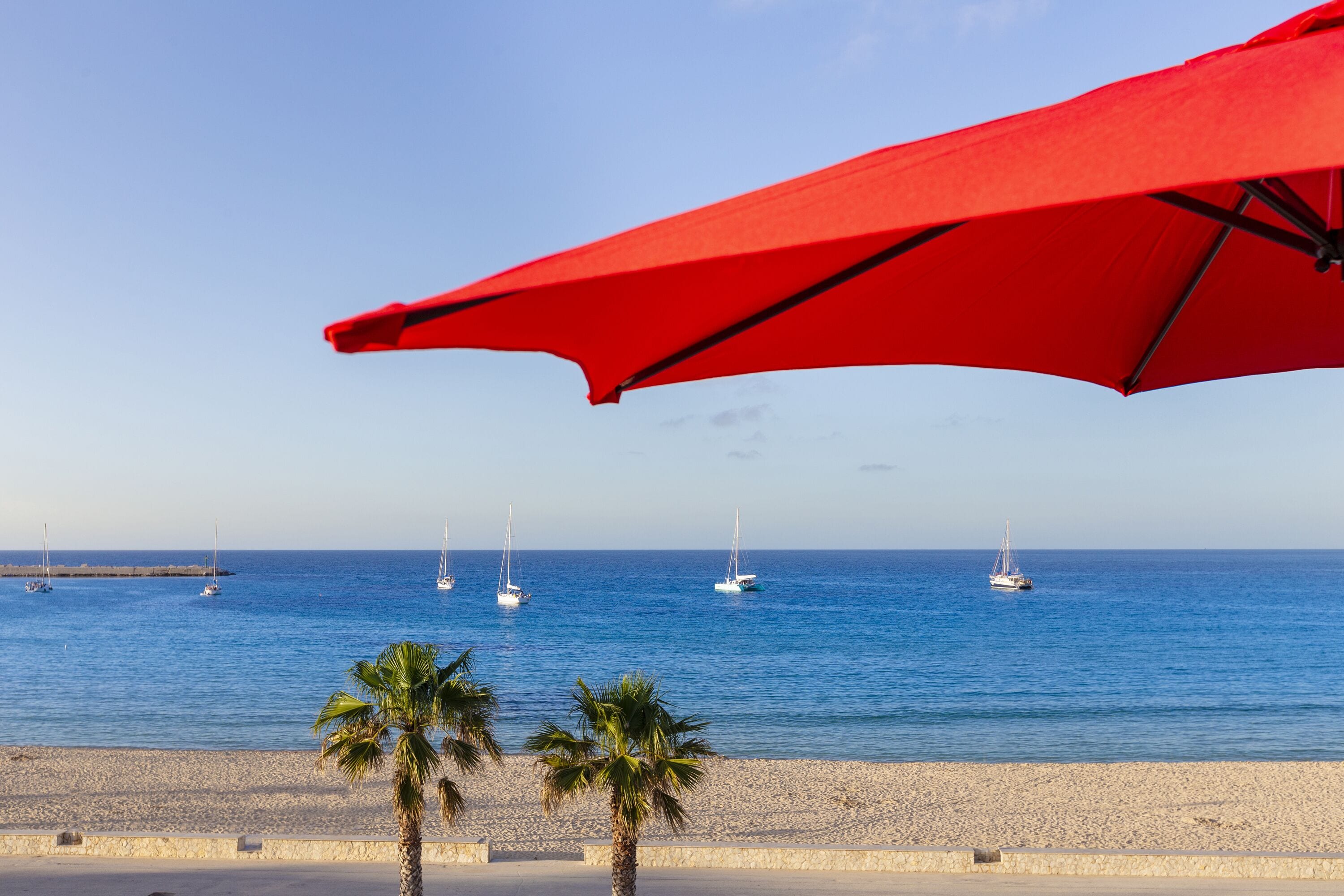 A beach with a red umbrella, palm trees, and sailboats on the water.