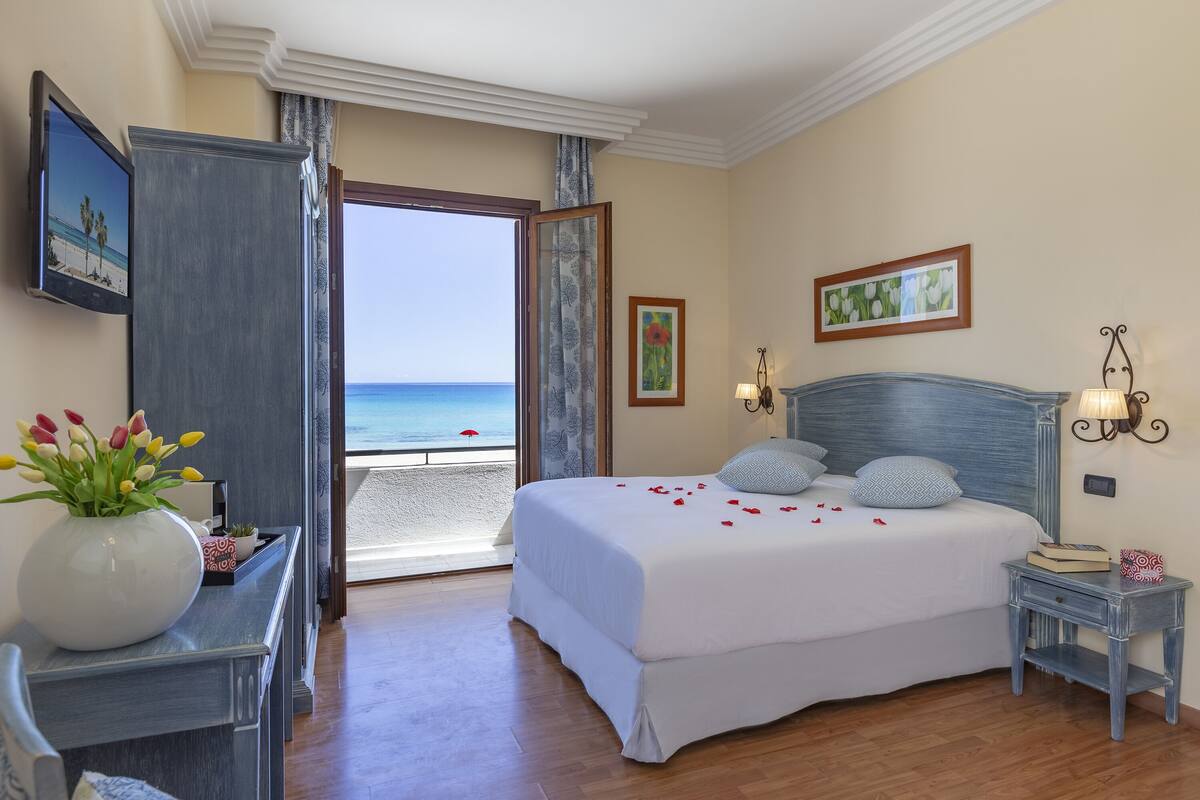 Een hotelkamer met een groot bed, een bureau met een bloevas, uitzicht op het strand en kunstwerken aan de wanden.