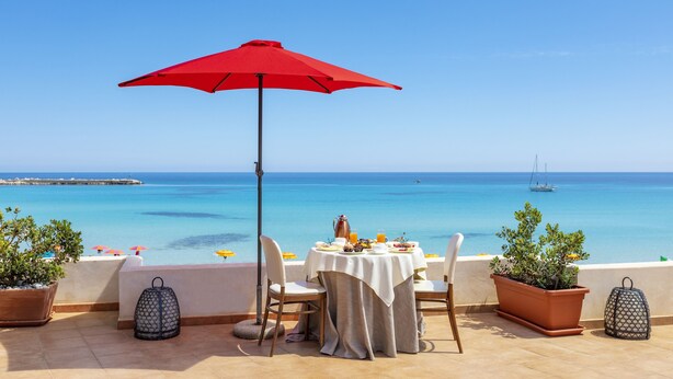 Een terras met een gedekte tafel voor twee, met uitzicht op een heldere blauwe zee en een pier.