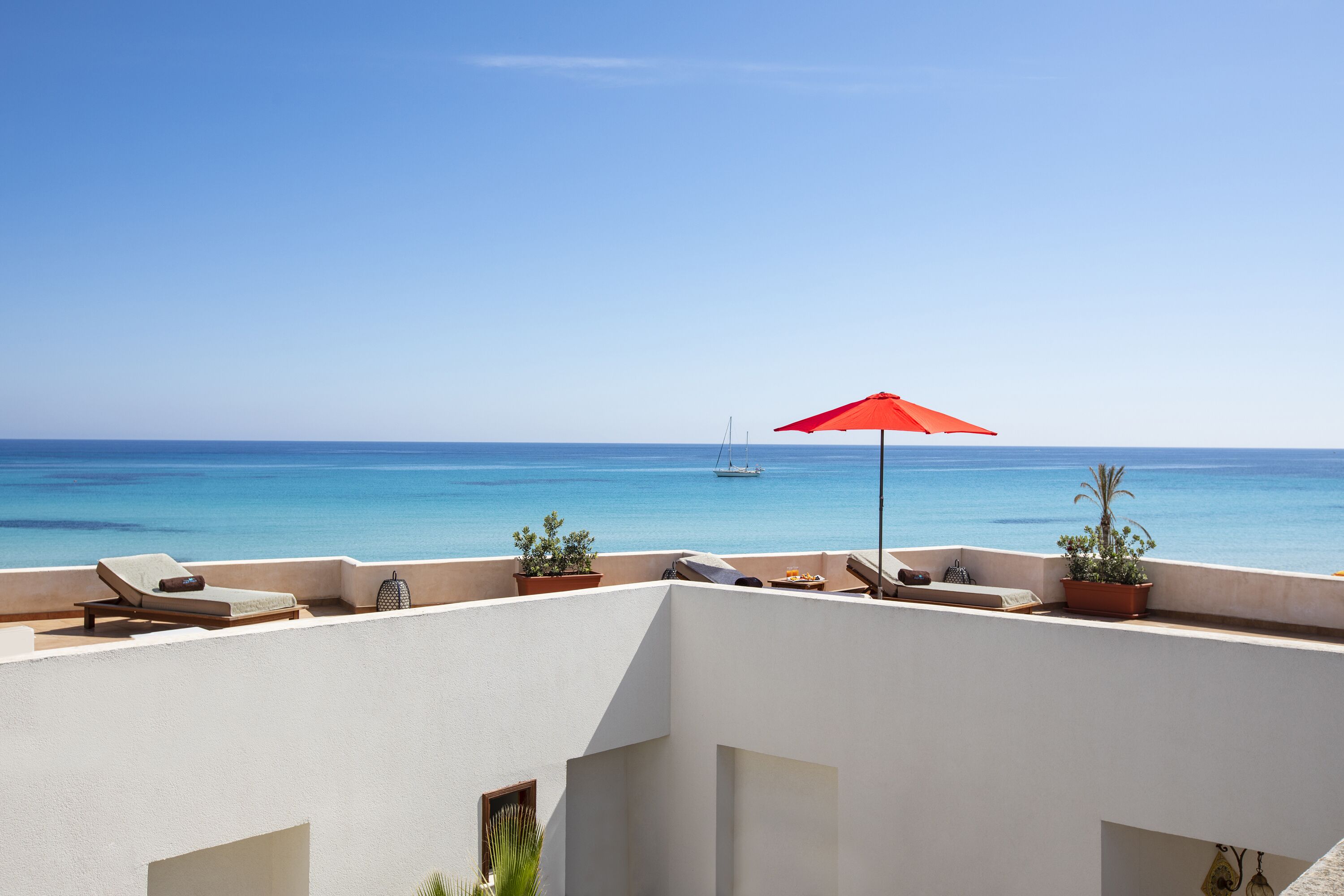 Eine Dachterrasse mit rotem Sonnenschirm, Liegestühlen, Topfpflanzen und Blick auf das Meer sowie ein Segelboot.
