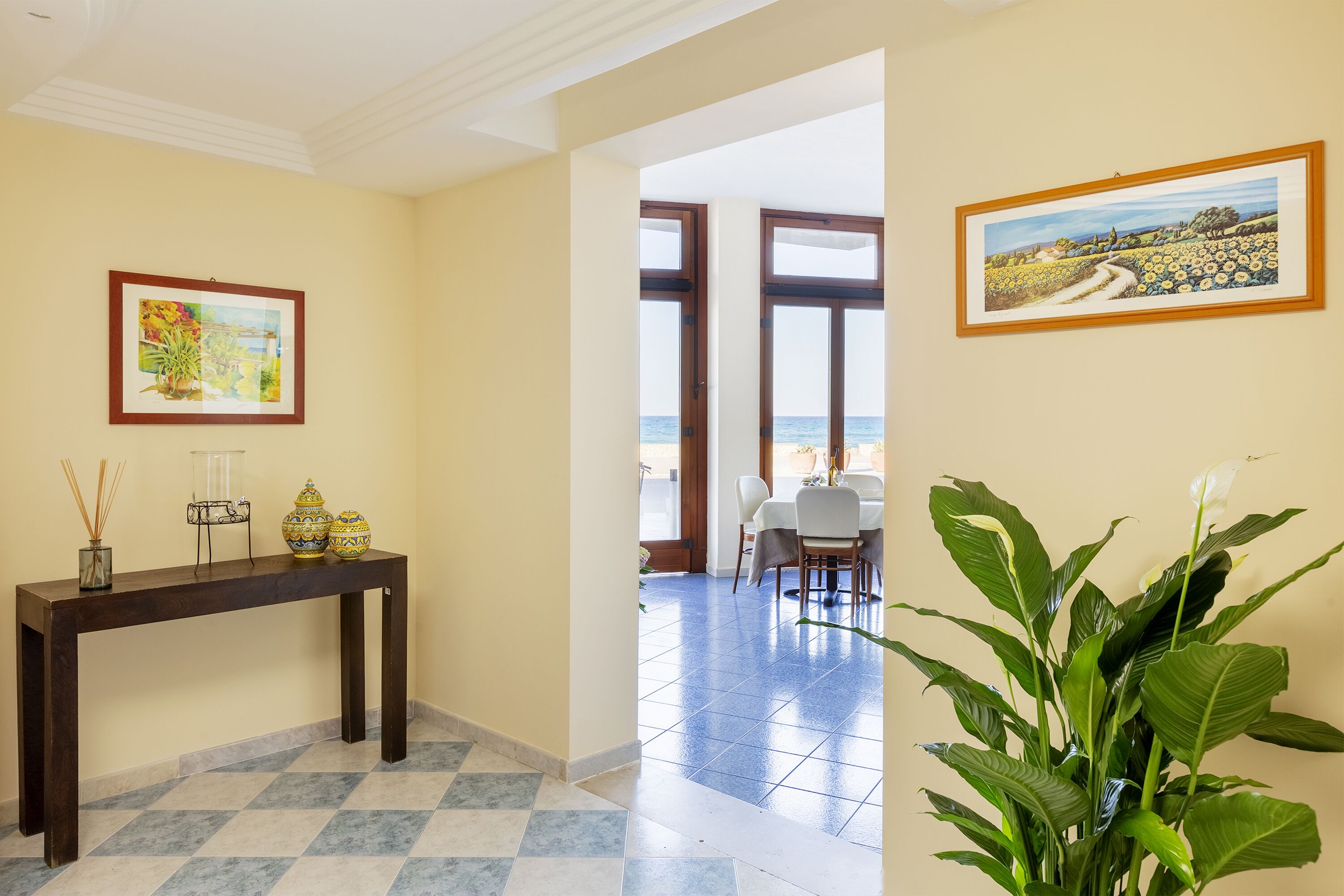 Una hall d'hotel con pavimento a scacchi, un tavolo consolle in legno con oggetti decorativi, un quadro incorniciato e vista sul mare attraverso ampie finestre.