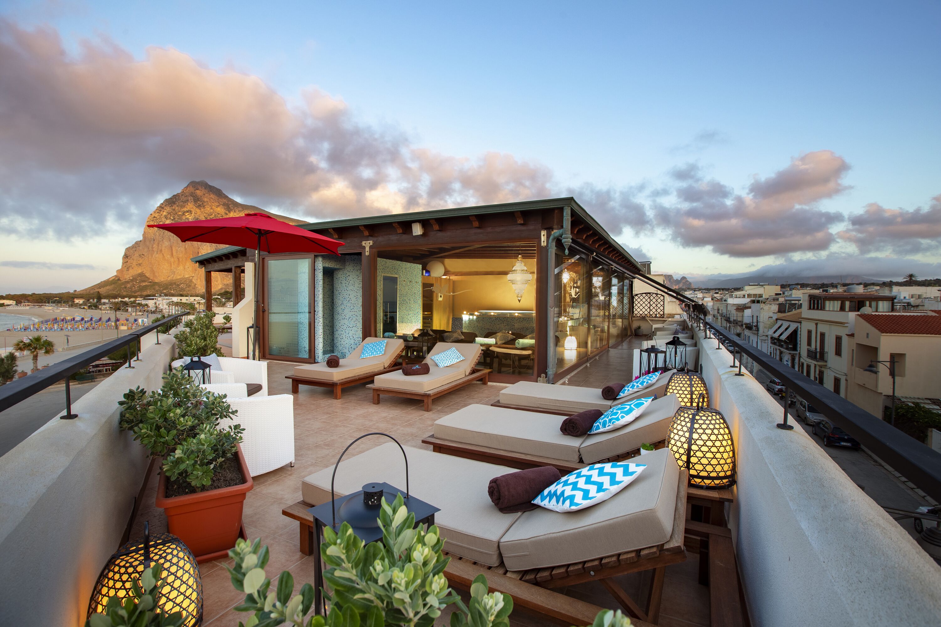 A rooftop terrace with lounge chairs, a red umbrella, and a view of a mountain and beach.