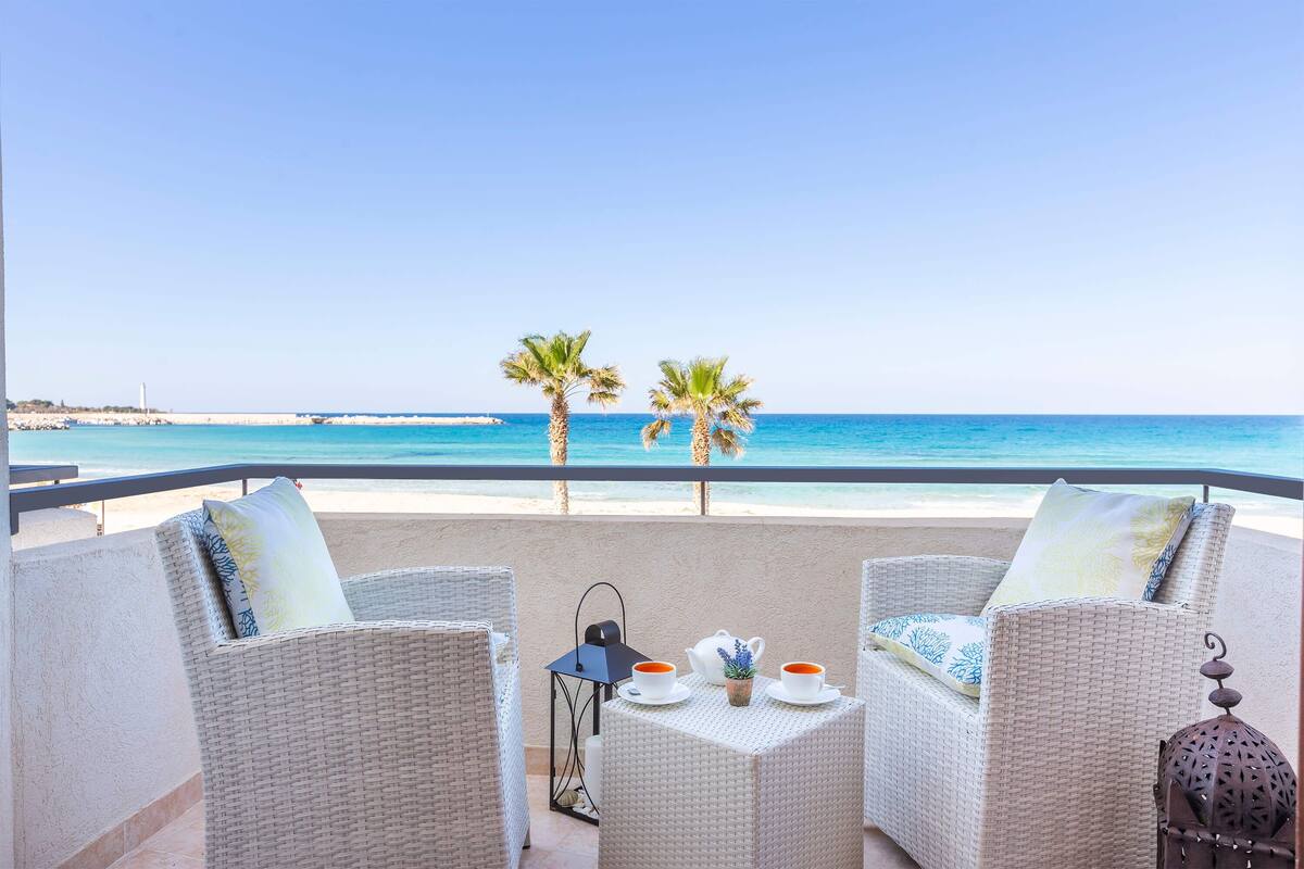 Een balkon met rieten meubilair, een tafel met theepotten en kopjes, en uitzicht op het strand en de oceaan.