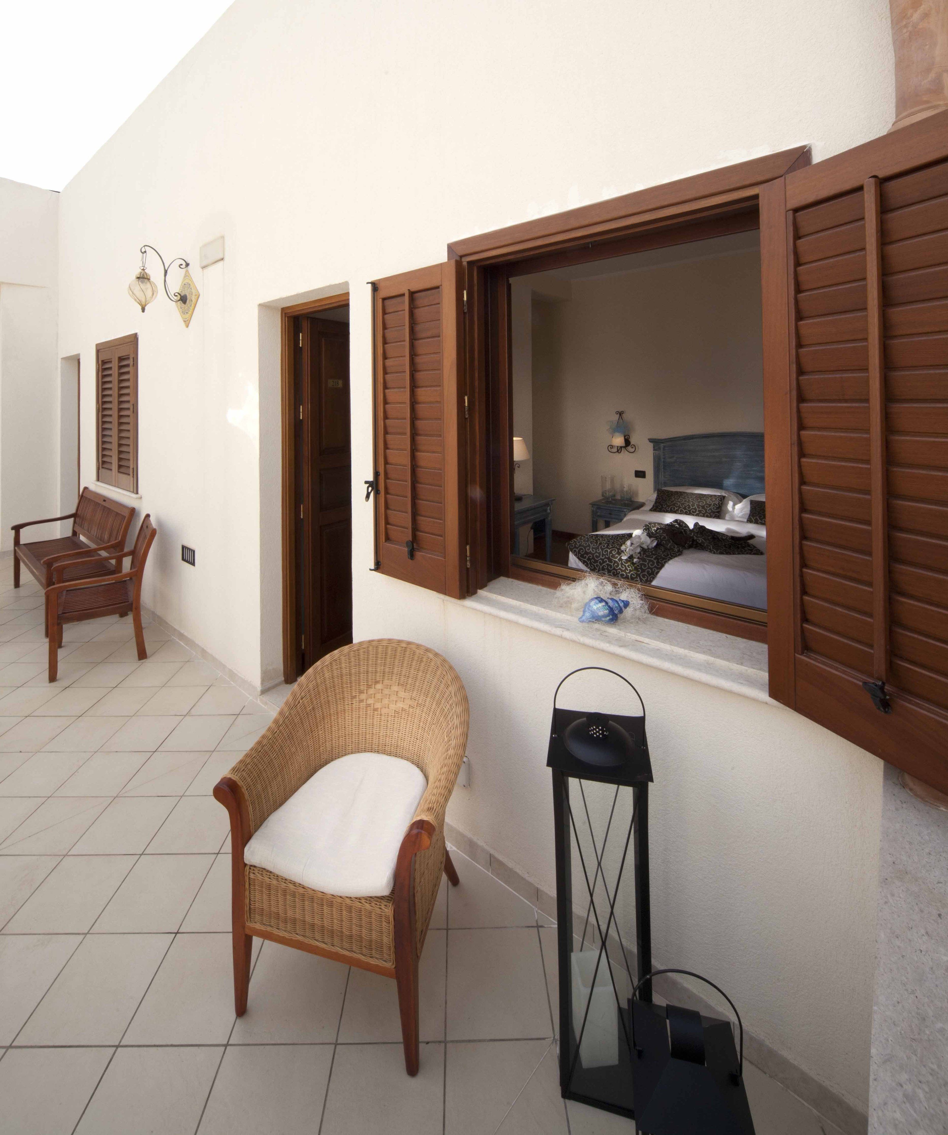 A room with a wicker chair, a bed with a headboard, a window with wooden shutters, and a small table with a lamp.