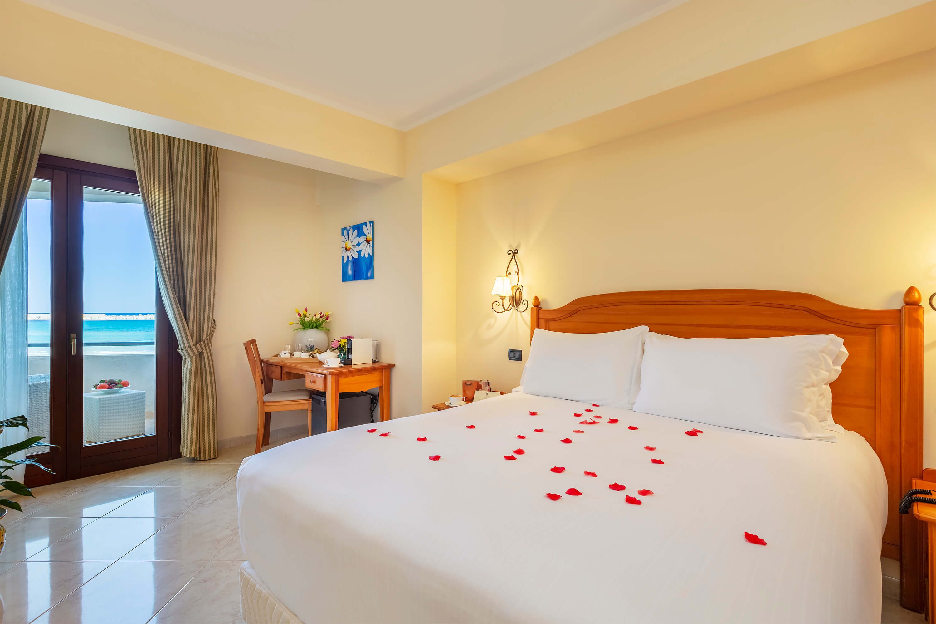 A hotel room with a bed, a desk, a chair, a nightstand, a lamp, and a view of the ocean through a sliding door.