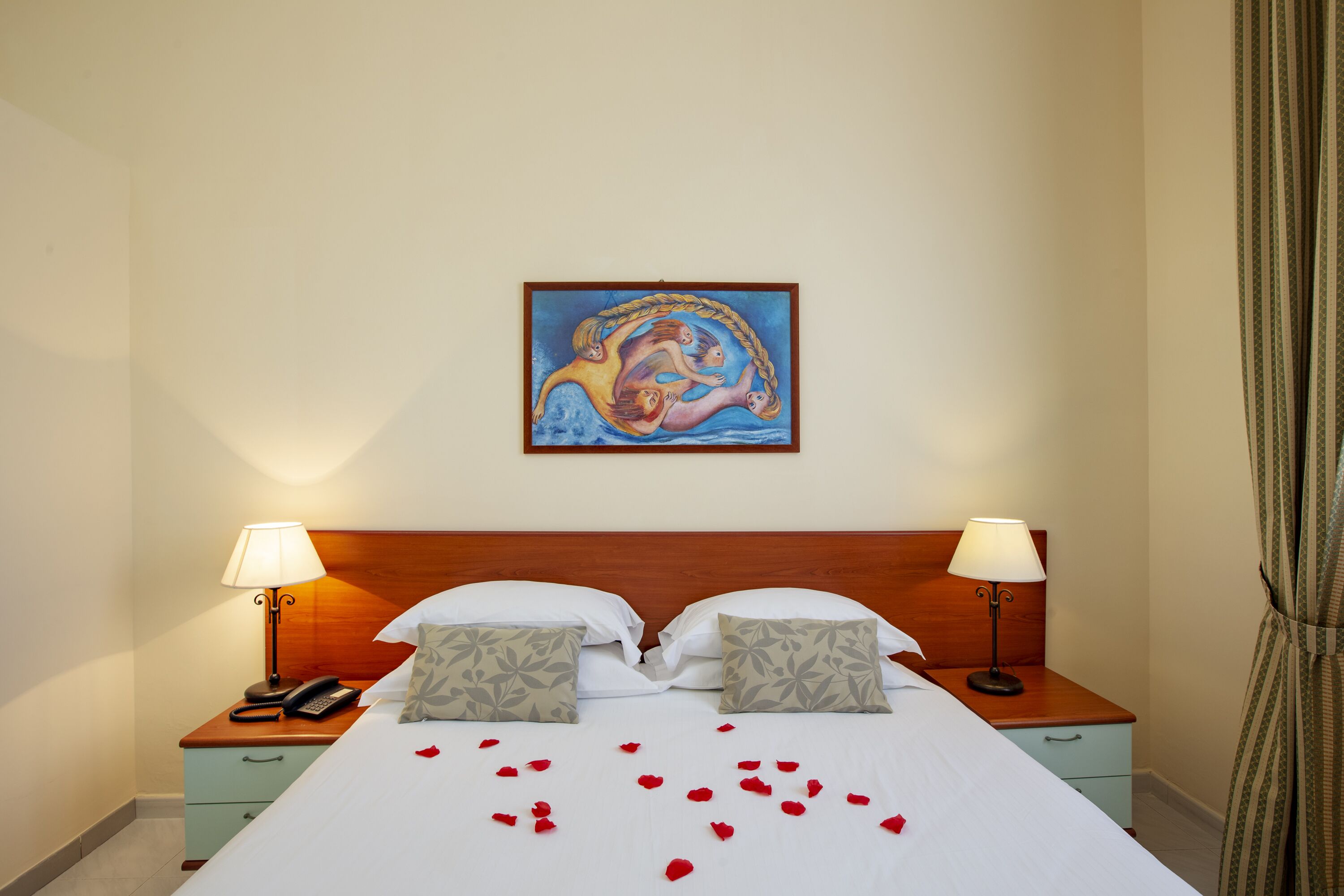 A hotel room with a bed, two bedside tables with lamps, a painting on the wall, and rose petals on the bed.