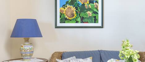 A living room with a wicker sofa, a blue armchair, a small round table with a lamp, and a framed picture of sunflowers on the wall.