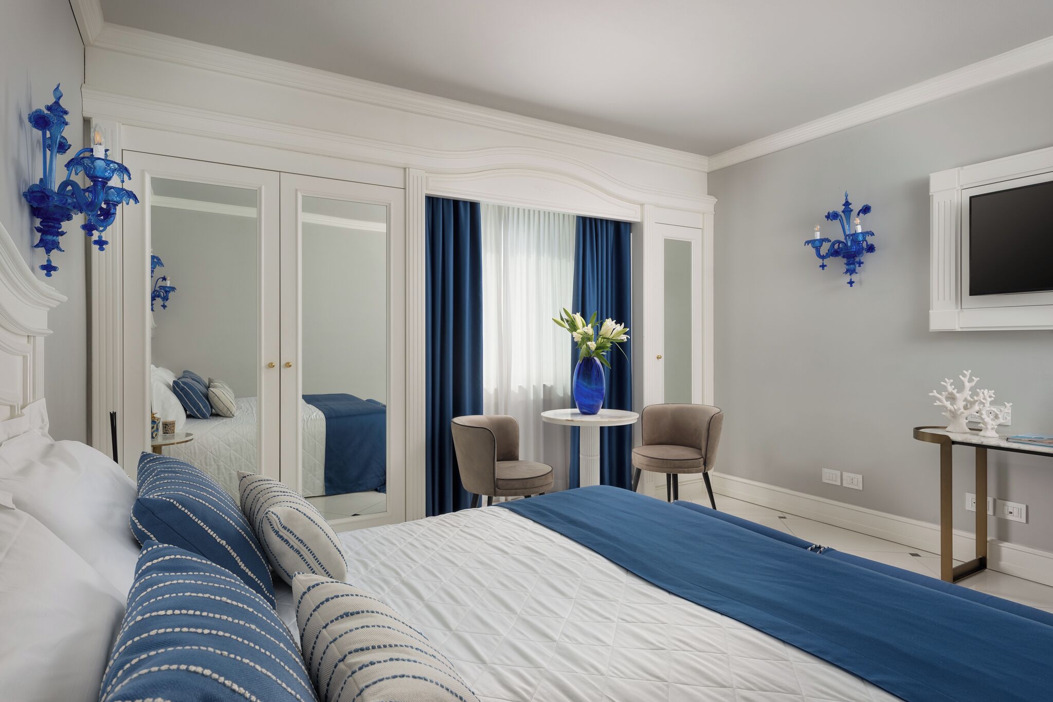 Quarto de hotel com uma cama, uma mesinha, duas cadeiras, uma televisão e um espelho.