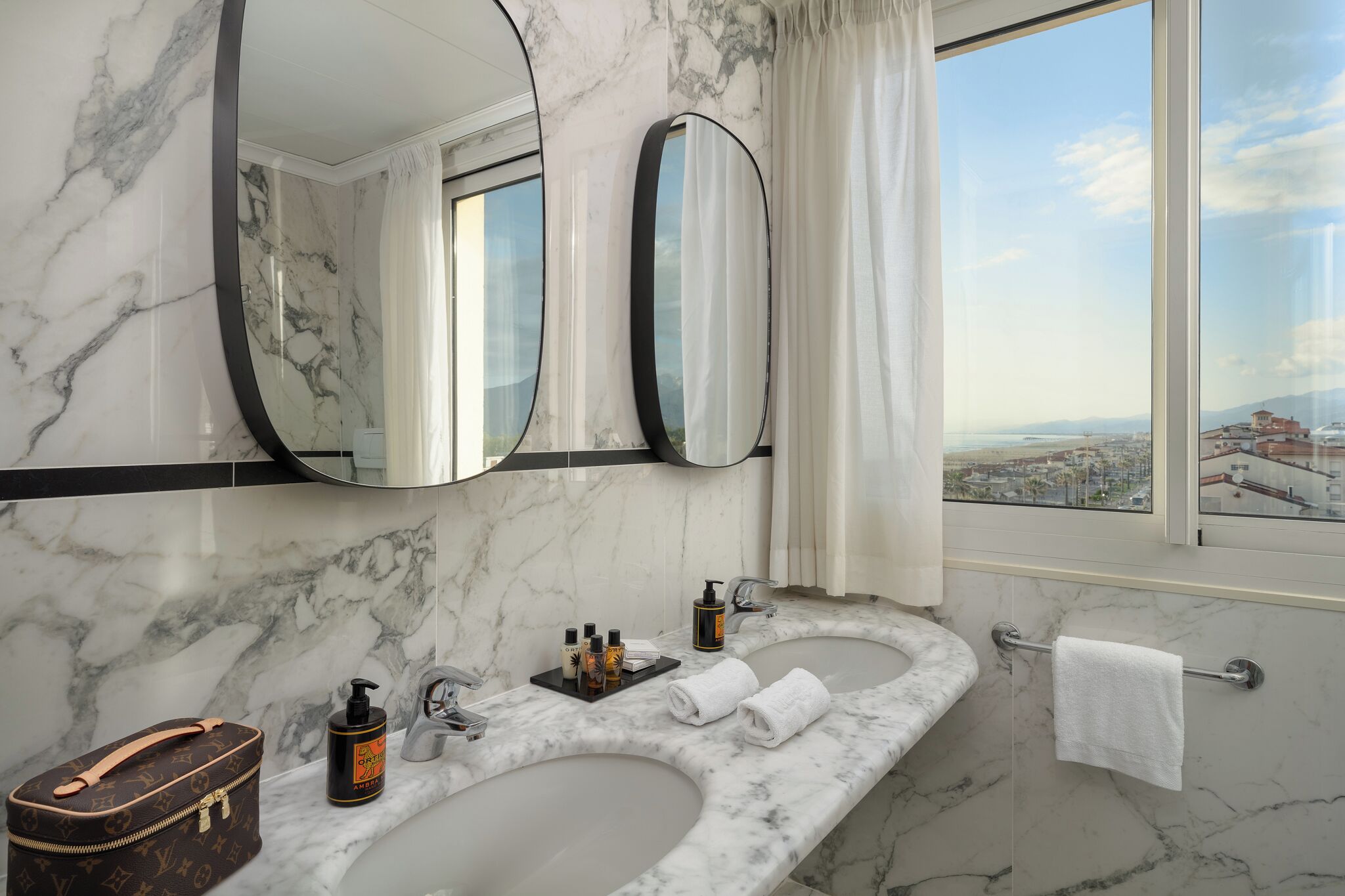 Une salle de bain aux murs recouverts de marbre, dotée de deux lavabos, d’un grand miroir et offrant une vue sur le paysage urbain par une fenêtre.