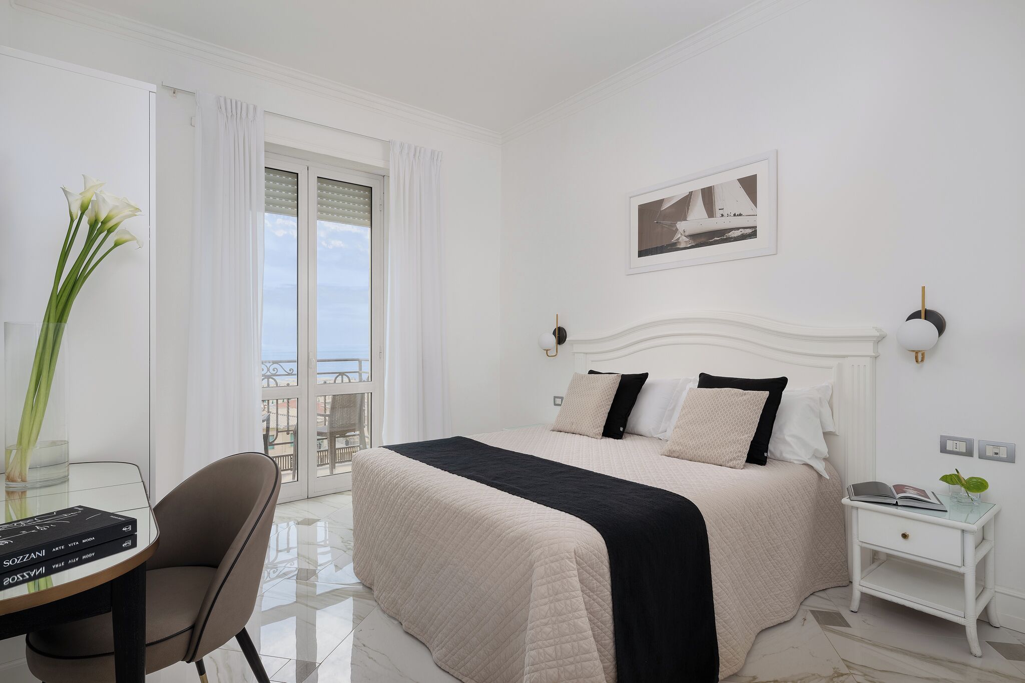 A hotel room with a bed, bedside table, a chair, a desk, a window with curtains, and a balcony.