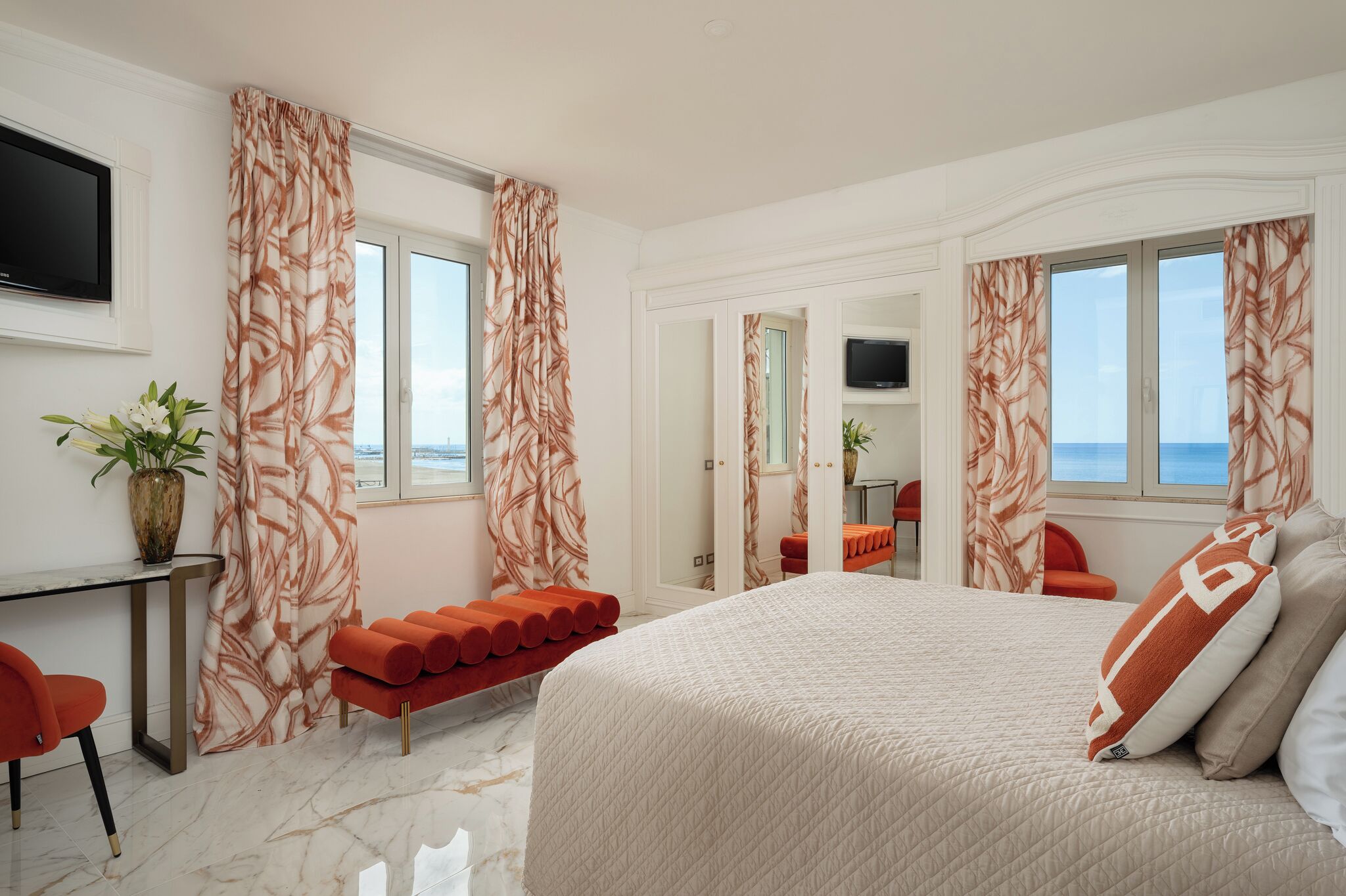 Habitación de hotel con cama, escritorio, silla, banco, televisor y vista al mar.