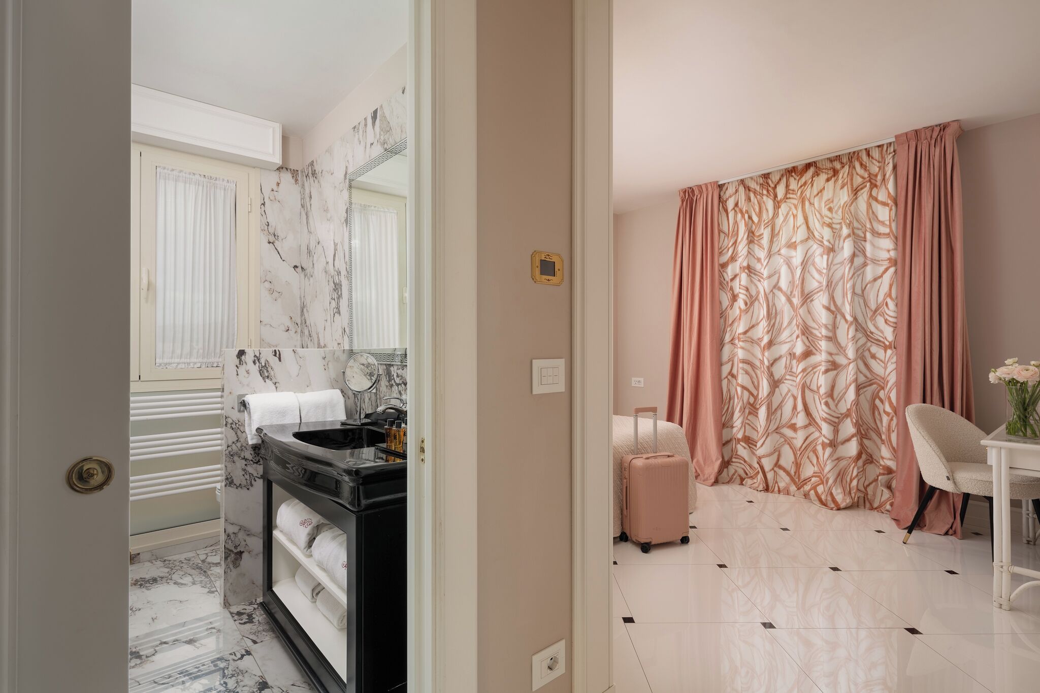 A hotel room with a marble bathroom, a bed with a pink suitcase, a white chair, and a table with a vase.