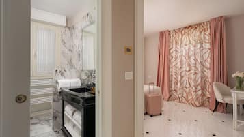 Une chambre d’hôtel avec une salle de bain en marbre, un lit avec une valise rose, une chaise blanche et une table avec un vase.